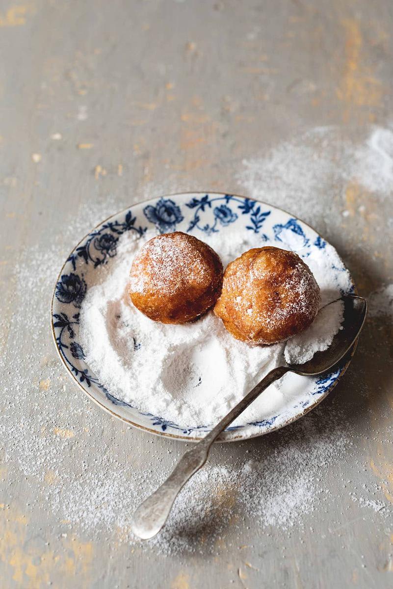 En una superficie gris está un plato con estampado azul. En él hay dos buñuelos de viento casero junto con mucho azúcar glas.