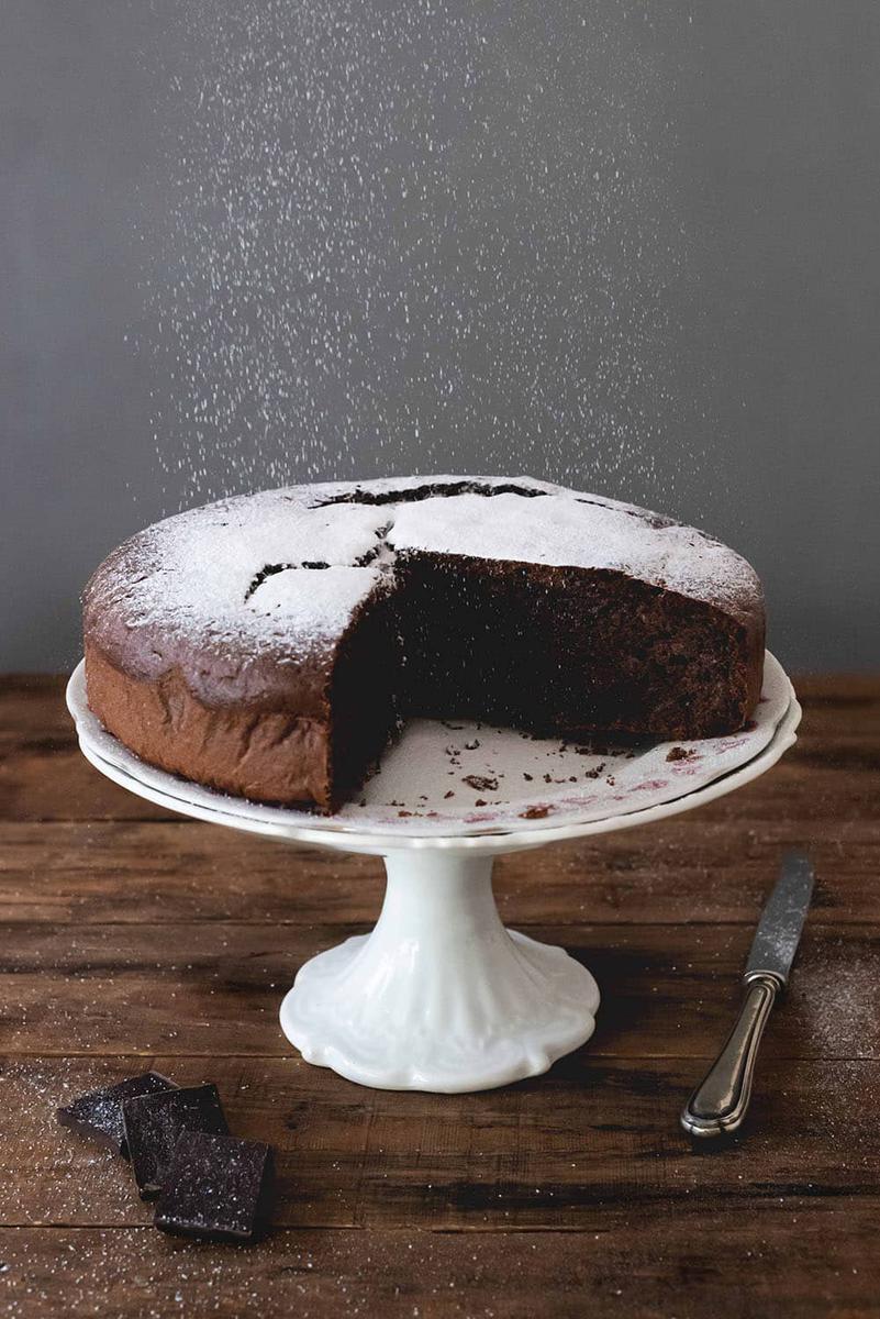 En una mesa de madera está el bizcocho de chocolate con aceite de oliva cortado encima de un porta tarta. También se ve que están espolvoreando con azúcar glas.