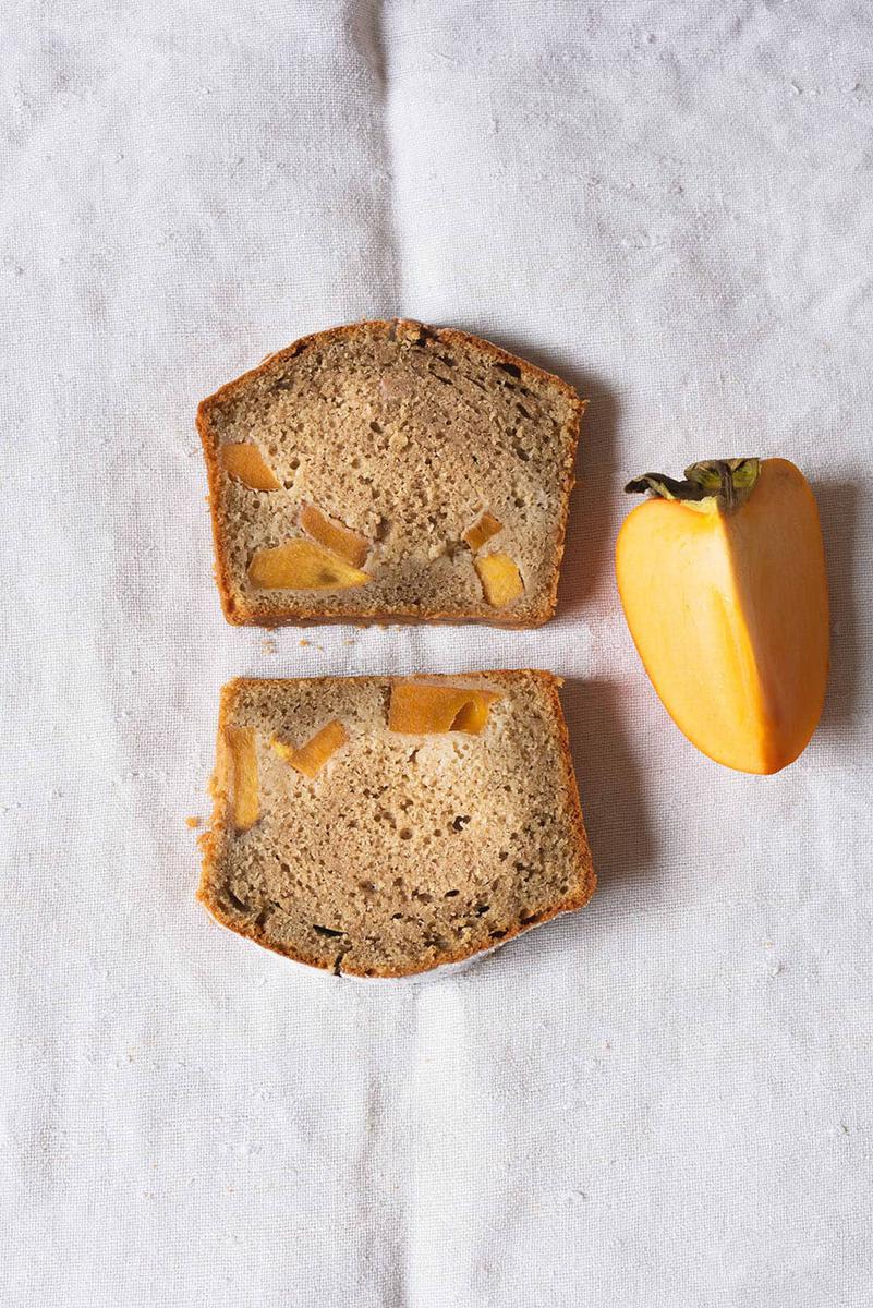En una mesa cubierta con un mantel blanco, están dos rodajas de caquis de bizcocho. Al lado hay un trozo de caqui.