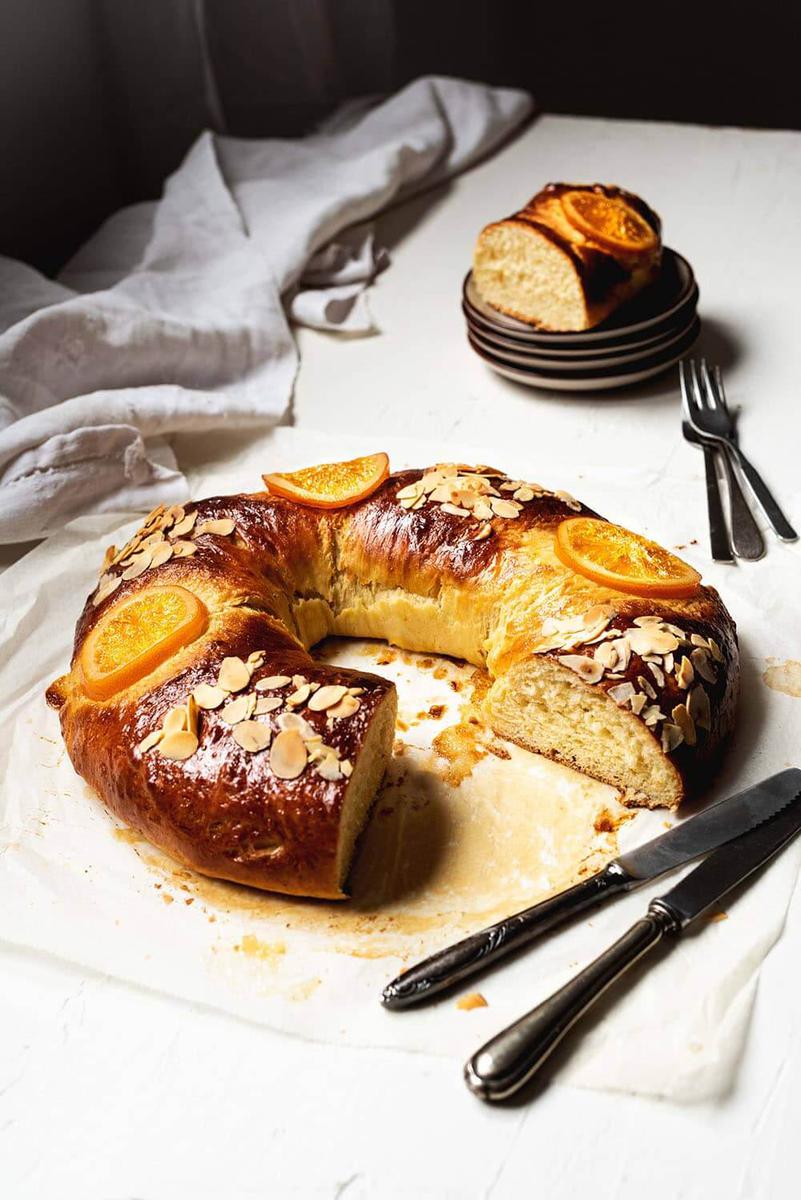 En una superficie blanca está un roscón de reyes tradicional de la abuela ya cortado. Se ve muy esponjoso. Alrededor hay cubiertos y unos platos blancos.