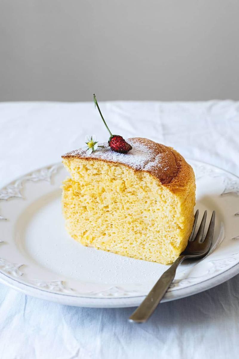En una mesa cubierta con mantel blanco hay un plato blanco. Encima del plato está una ración de tarta de queso japonesa esponjosa, decorada con una fresa y una flor de fresa. . Se ven un tenedor también.