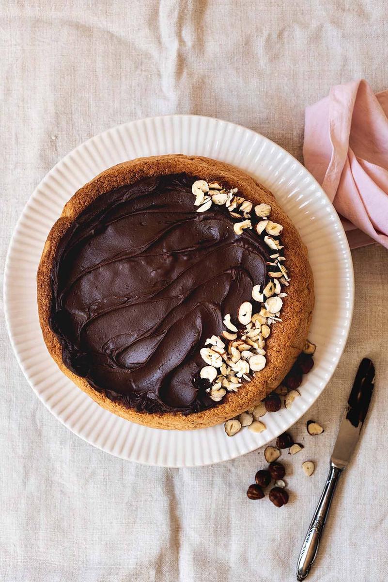 La mesa está cubierta con un mantel blanco. En ello está el bizcocho esponjoso de avellanas y chocolate encima de un plato blanco. Está sin cortar.