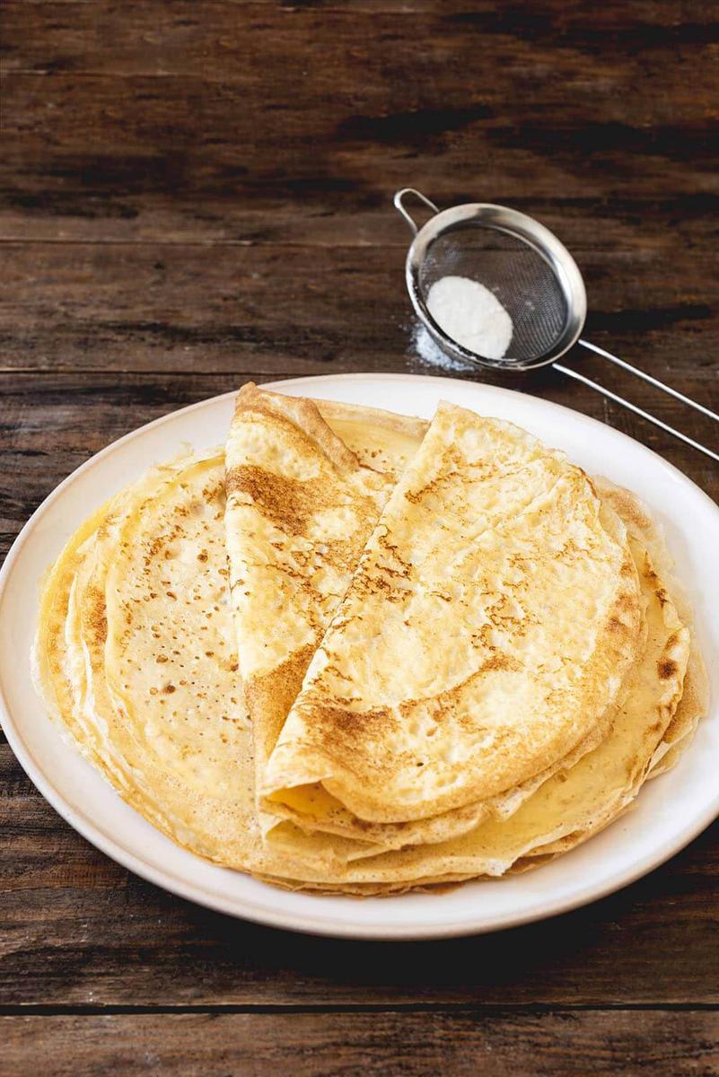 En la mesa de madera hay un plato. En el plato hay varios frixuelos asturianos tradicionales. Alrededor hay un tamiz para espolvorear con azúcar glas los frixuelos. 