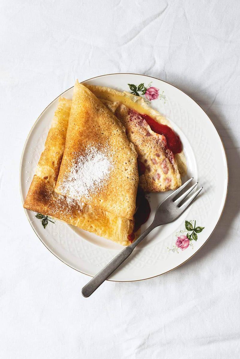 En la mesa cubierta con un mantel blanco hay un plato. En el plato hay dos frixuelos asturianos tradicionales rellenos con mermelada de fresas.