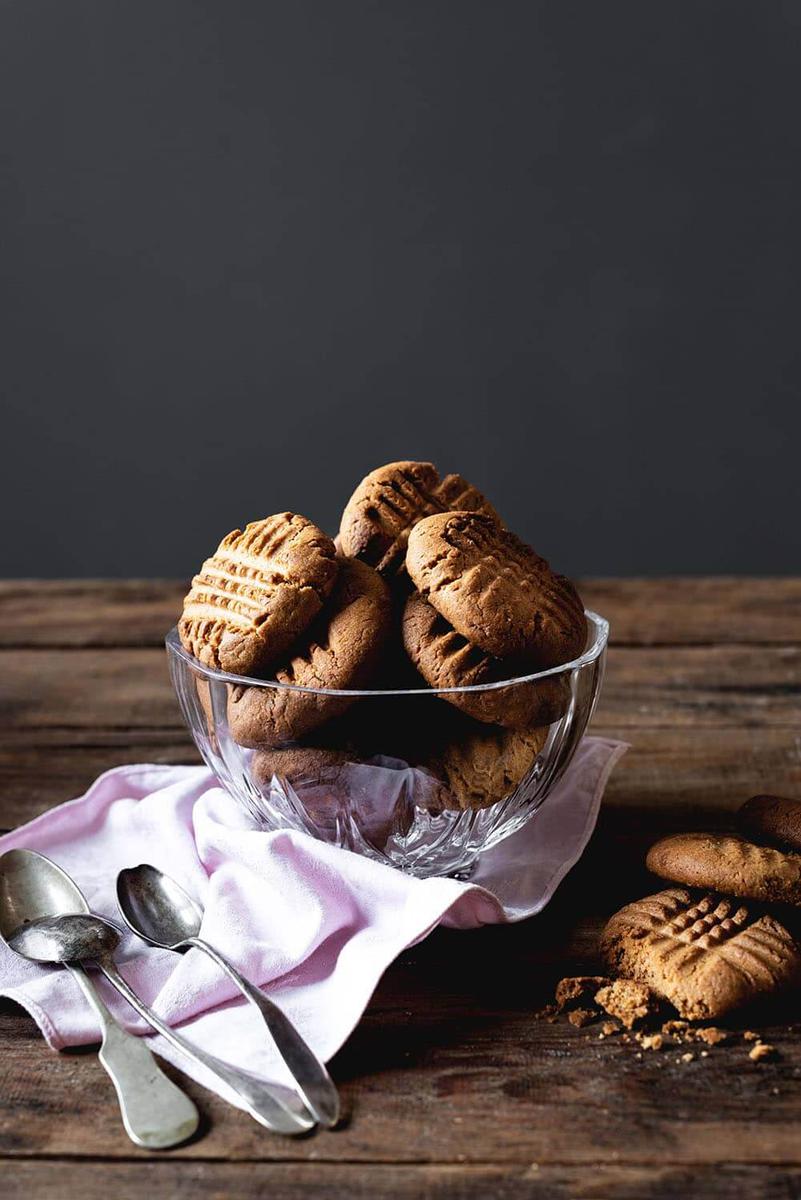 En la mesa de madera hay un recipiente transparente lleno de galletas de mantequilla de cacahuete caseras. Alrededor hay un paño rosa y cubiertos.