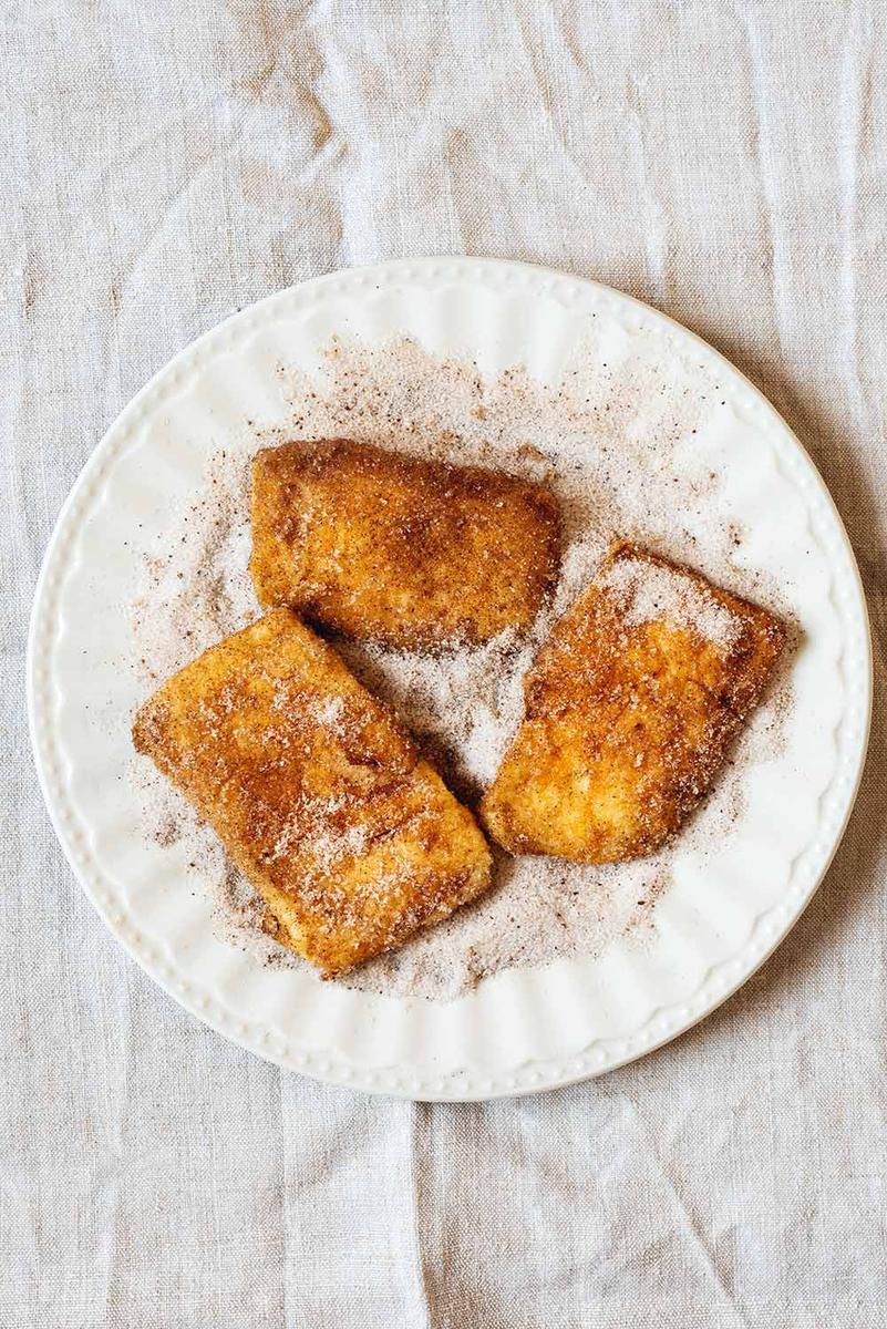 En una mesa cubierta con mantel blanco hay hay un plato blanco lleno de una mezcla de azúcar y canela. En él hay tres raciones de leche frita casera con maicena. Se ven muy cremosas.