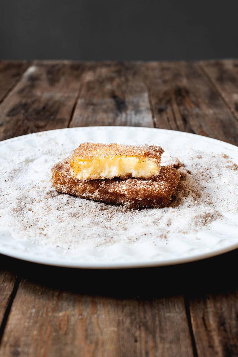En una mesa de madera hay un plato blanco lleno de una mezcla de azúcar y canela. En él hay dos raciones de leche frita casera con maicena. Se ven muy cremosas.