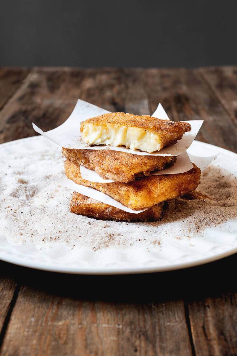 En una mesa de madera hay un plato blanco lleno de una mezcla de azúcar y canela. En él hay cuatro raciones de leche frita casera con maicena. Es como si fuera una torre. Entre las capas de leche fruta hay papel de horno. Se ven muy cremosas.