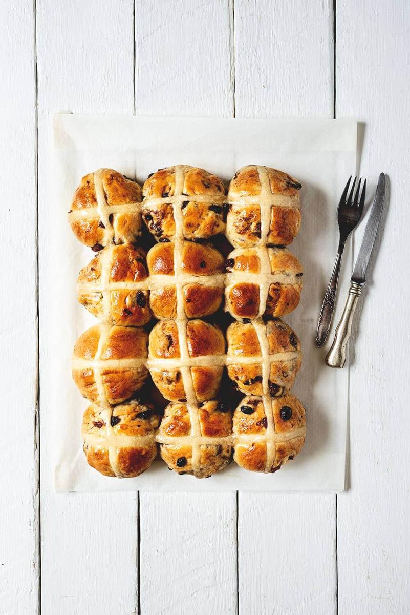 En una mesa blanca hay un trozo de papel de horno. Encima hay doce Hot Cross Buns. Al lado hay unos cubiertos.