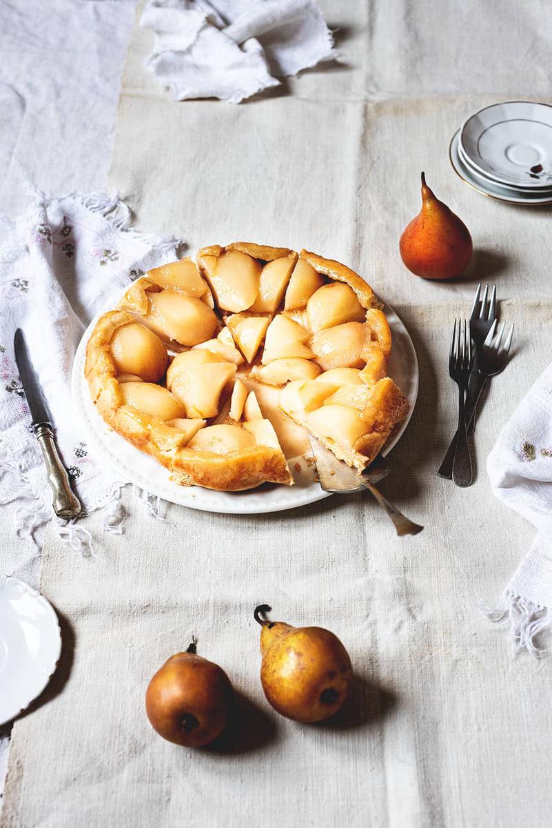 Una mesa está cubierta con un mantel blanco. Encima está la Tarta Tatín de peras. Ya está cortada. Un toro está en una pala de tarta. Alrededor de la tarta hay varios manteles blancos cubiertos y platos. También hay dos peras Conferencia.