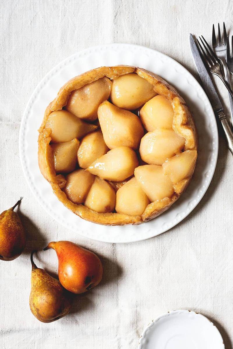 Una mesa está cubierta con un mantel blanco. Encima está la Tarta Tatín de peras. Alrededor de la tarta hay cubiertos y peras Conferencia.