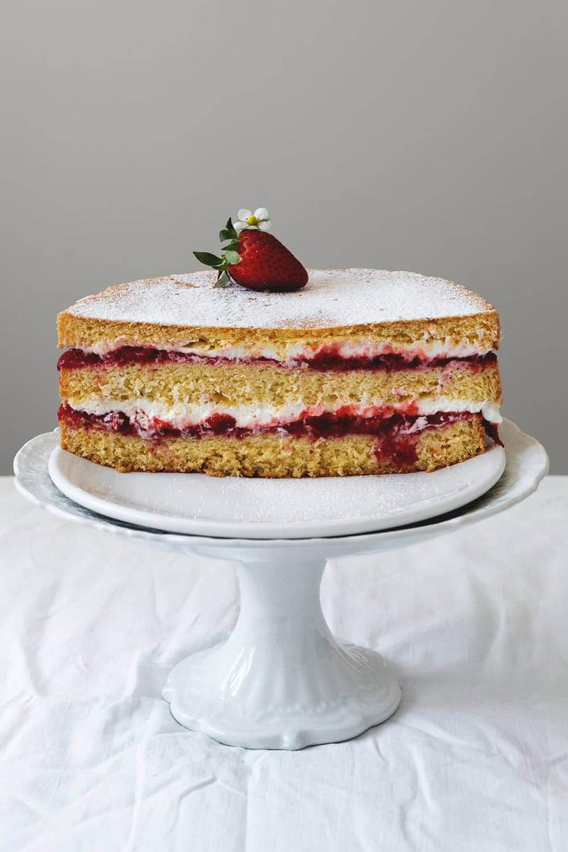 En una mesa cubierta con mantel blanco está una portatarta blanca. En ella está la tarta de fresas con bizcocho genovés cortado en la mitad. 