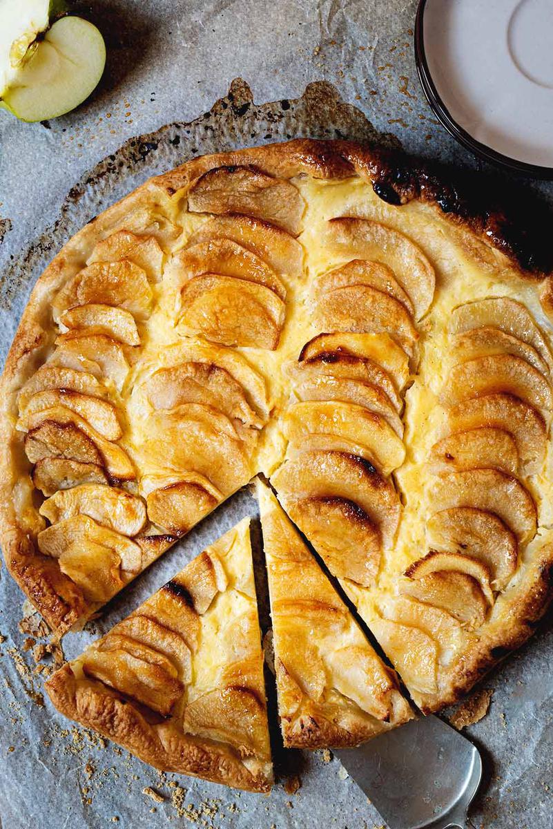 En una mesa de madera está puesto el papel de horno y encima está la tarta de manzana, crema pastelera y hojaldre. Está brillante y ya está cortada. Alrededor hay manzana fresca y unos platos de color marrón.