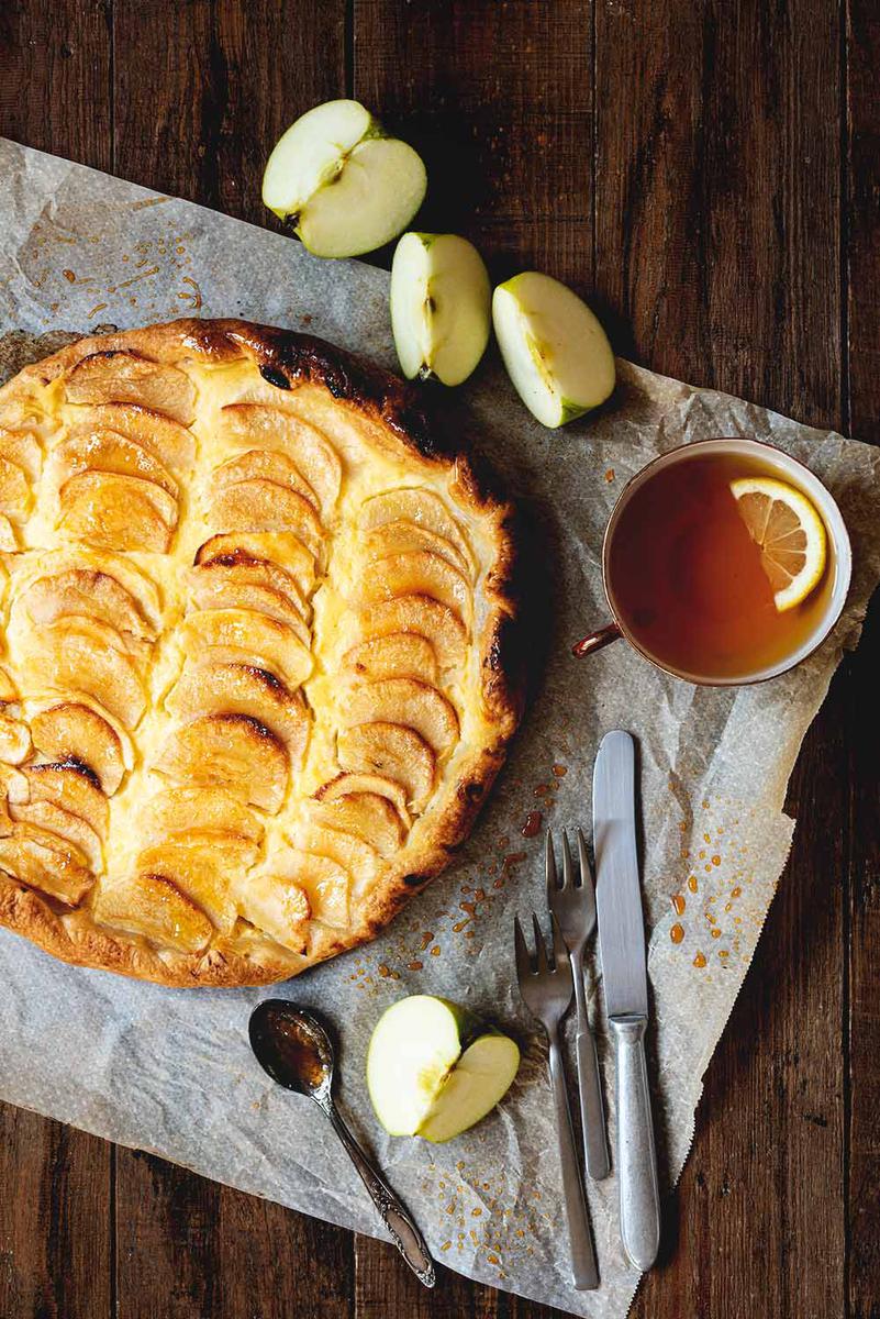 En una mesa de madera está puesto el papel de horno y encima está la tarta de manzana, crema pastelera y hojaldre. Está brillante. Alrededor hay una taza de té, cubiertos y unos gajos de manzanas frescas.