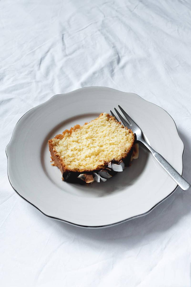 En una mesa cubierta con mantel blanco hay un plato blanco. Encima está una rodaja de bizcocho de coco de la abuela.