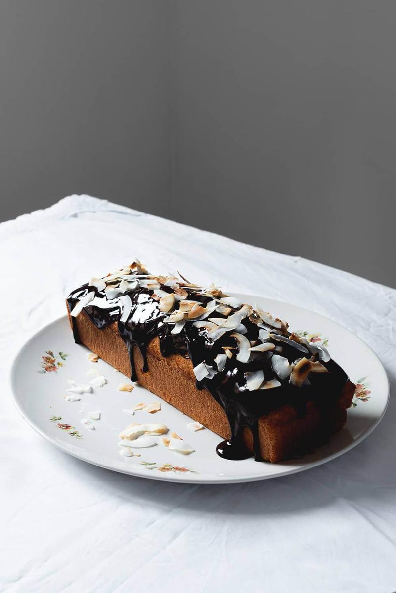 En una mesa cubierta con mantel blanco hay un plato grande y blanco. Encima está el bizcocho de coco de la abuela sin cortar. Se ve la salsa de chocolate y los chips de coco tostados encima.