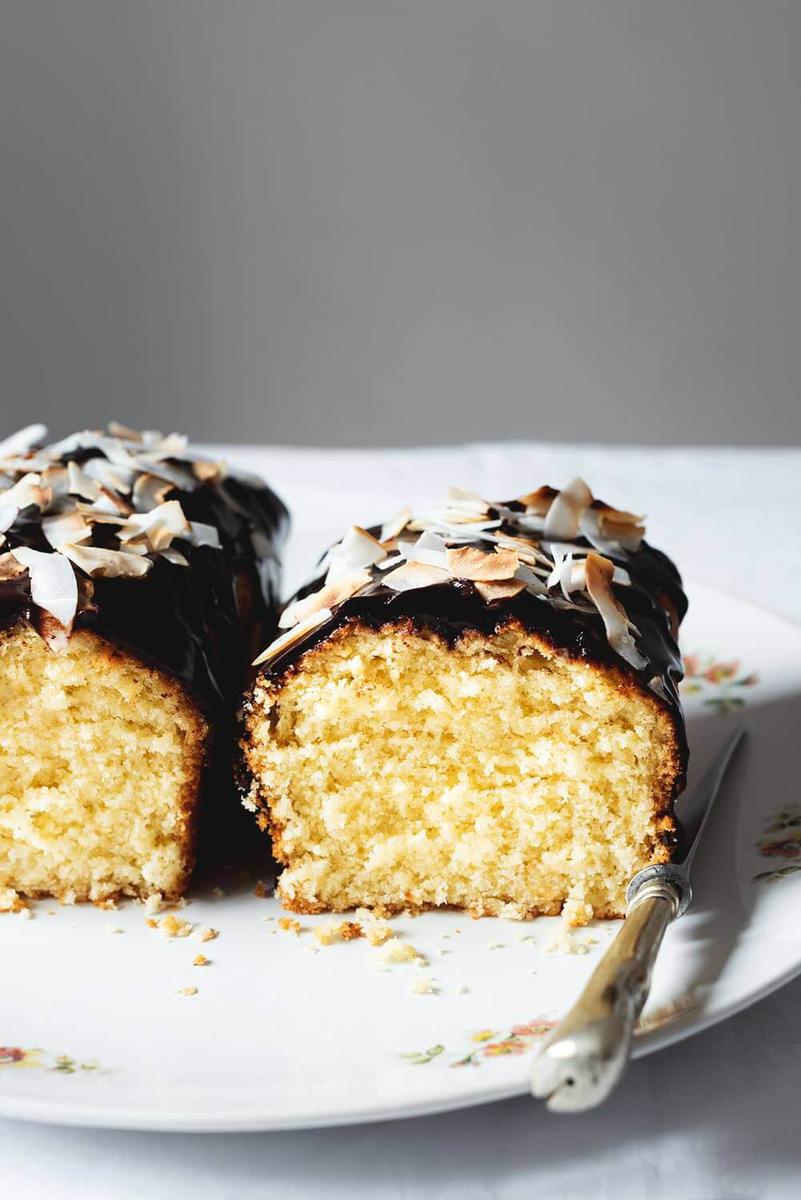 En una mesa cubierta con mantel blanco hay un plato grande y blanco. Encima están dos trozos de bizcocho de coco de la abuela Se ve esponjoso el interior, la salsa de chocolate y los chips de coco tostados encima.