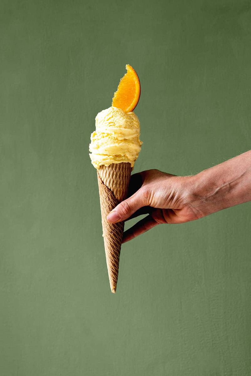 Delante de una pared verde una persona está sujetando un cucurucho lleno de helado de naranja casero. Como decoración hay un gajo de naranja encima de última bola de helado. 