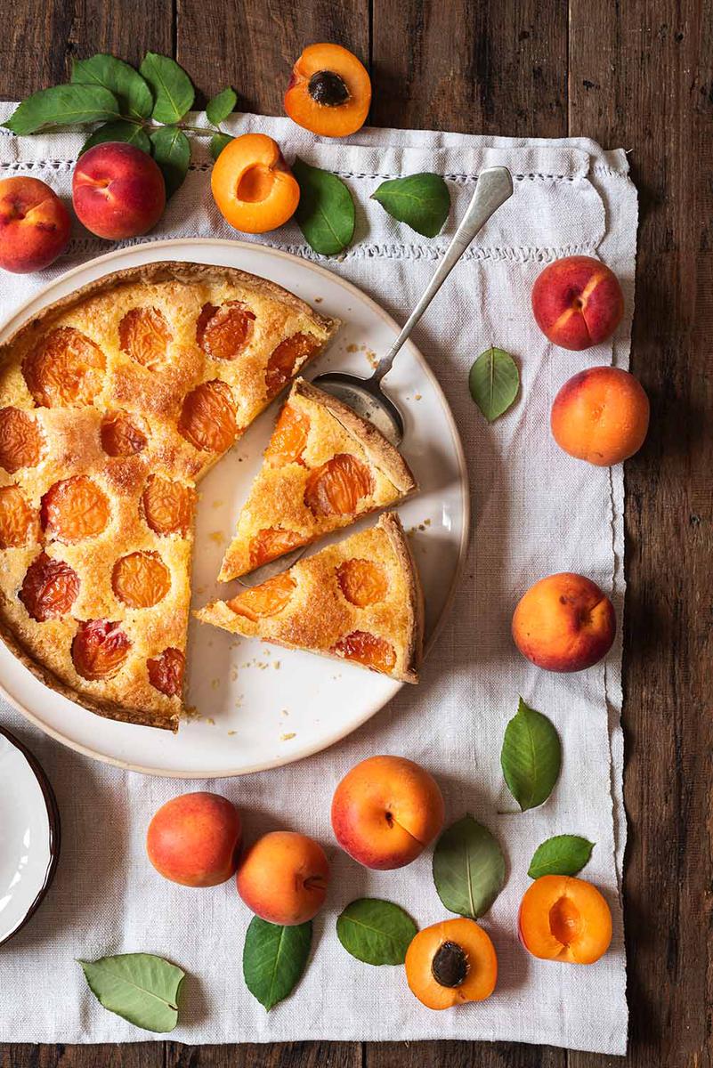 En una mesa de madera hay un paño de cocina blanca. Encima está la tarta de albaricoque con masa quebrada. Alrededor hay albaricoques frescos, un plato y hojas verdes. La tarta está cortada y abajo de una ración hay una pala de tarta.