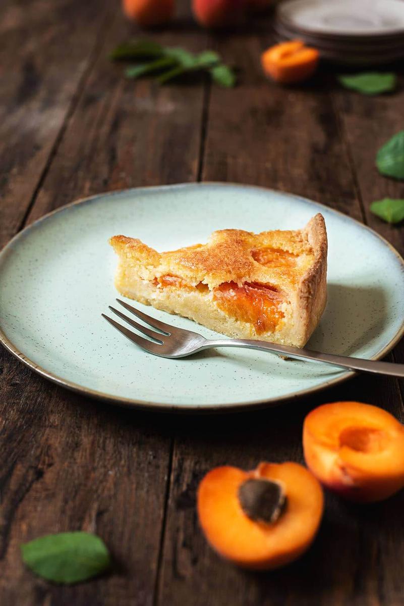 En una mesa de madera hay un plato de color turquesa. En el plato hay una ración de tarta de albaricoque con masa quebrada. Al lado del postre hay un tenedor. Alrededor del plato hay varios albaricoques frescos, hojas verdes y un par de platos.