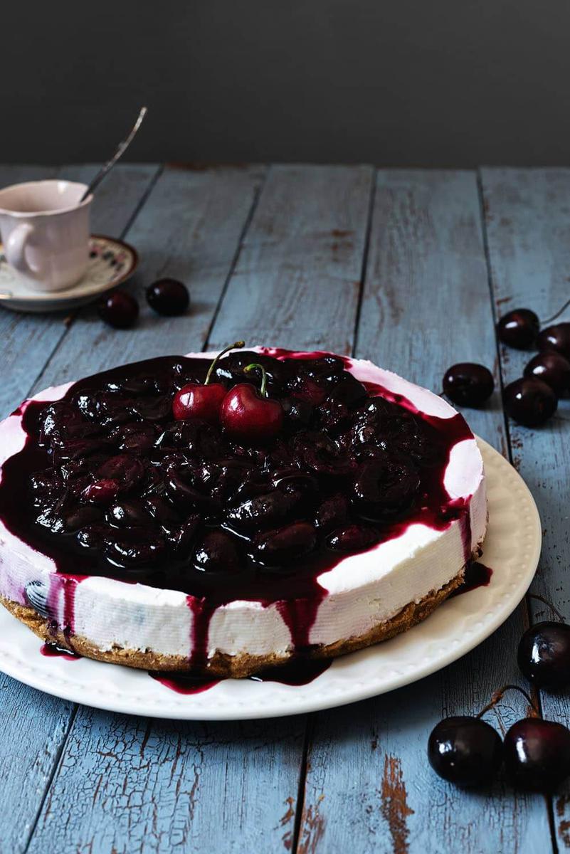 En una mesa azul hay un plato blanco. Encima está la tarta de queso con cerezas. Se ve que está muy jugoso por la salsa de cerezas. Alrededor del plato hay varias cerezas frescas y una taza con cubiertos en el interior.