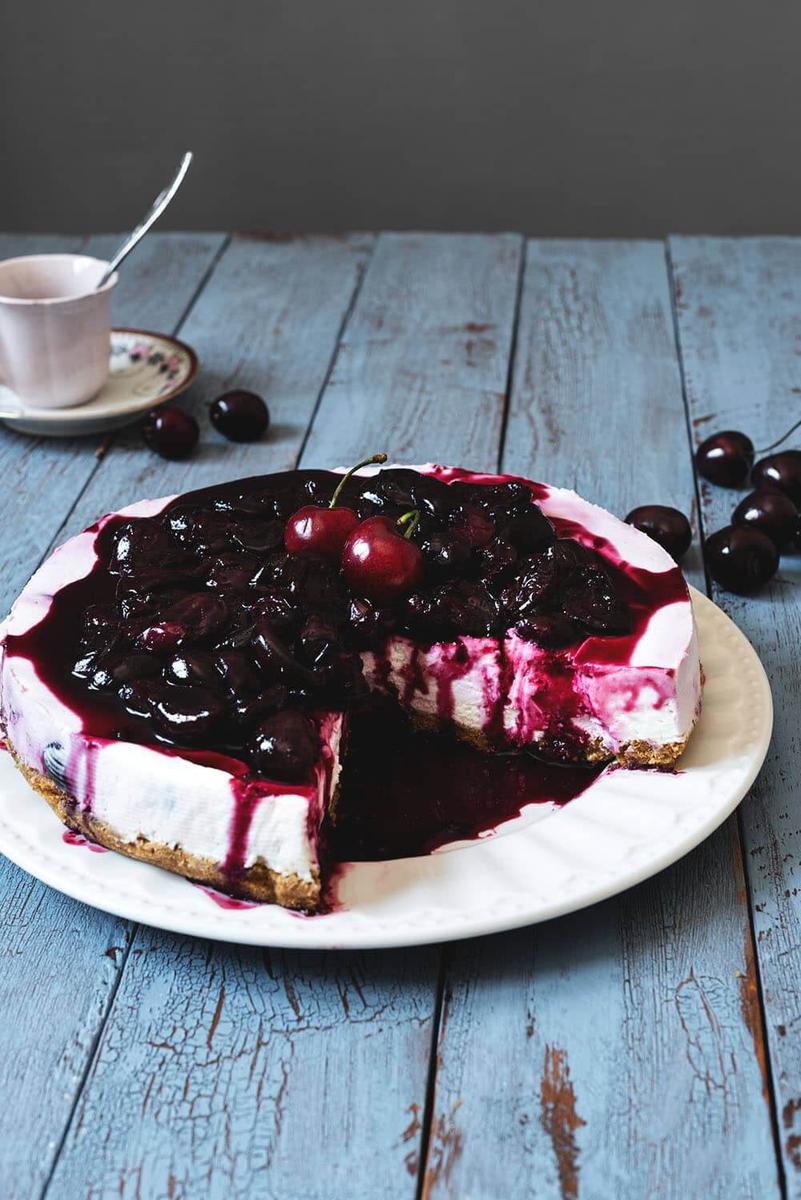 En una mesa azul hay un plato blanco. Encima está la tarta de queso con cerezas. Ya está cortado y se ve muy jugoso por la abundante cantidad de salsa de cerezas. Alrededor del plato hay varias cerezas frescas y una taza con cubiertos en el interior,