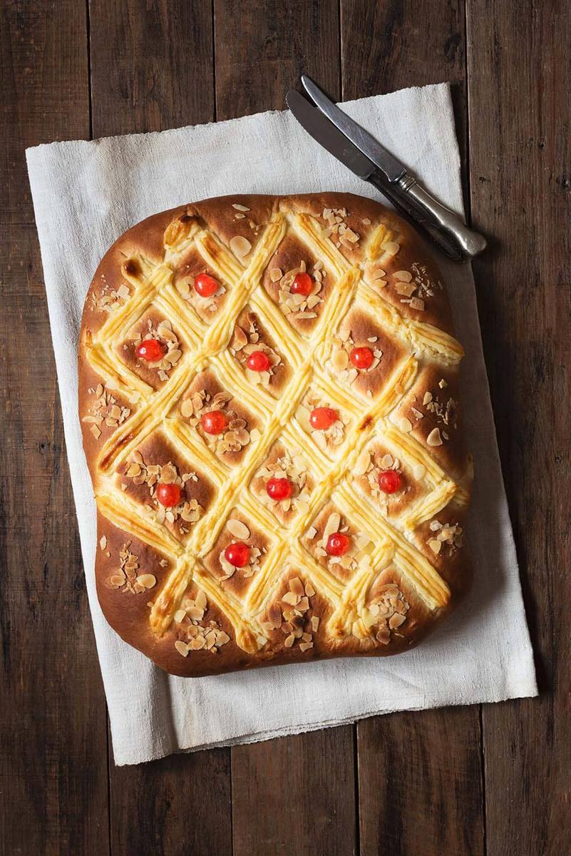 En una mesa de madera está puesto un paño de cocina. Encima está puesta la Coca de San Juan con crema pastelera. Al lado hay un cuchillo.