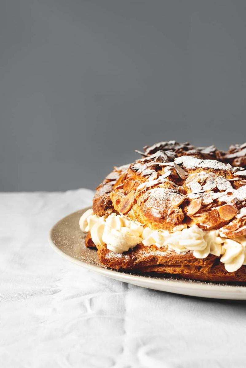 En una mesa cubierta con un mantel blanco hay un plato blanco. Encima está el Paris Brest. En la foto se ve el pastel muy cerca y solo se ve la mitad. Está espolvoreado con azúcar glas.