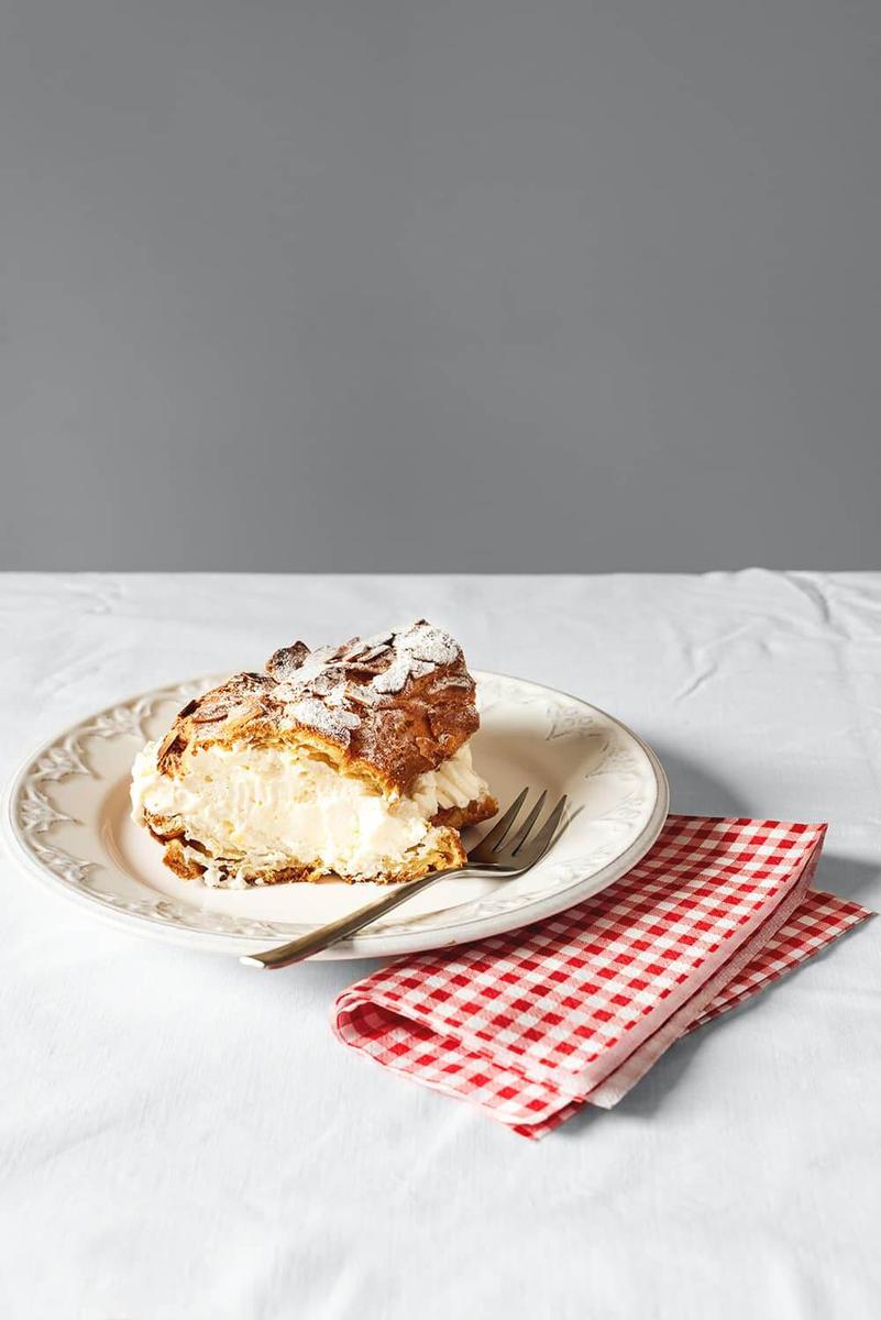 En una mesa cubierta con un mantel blanco hay un plato blanco. Encima hay una ración de Paris Brest. En el plato hay un tenedor. Al lado del plato hay una servilleta de cuadros de colores rojo y blanco.