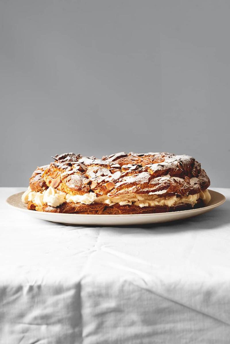 En una mesa cubierta con un mantel blanco hay un plato blanco. Encima está el Paris Brest. Está espolvoreado con azúcar glas.