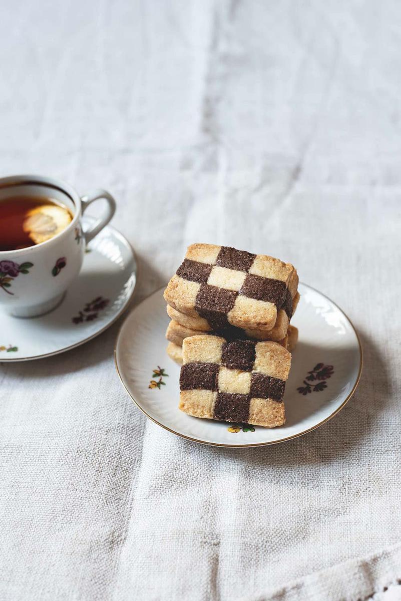 Una mesa está cubierta con un mantel blanco. Encima hay un plato pequeño con galletas de ajedrez. Al lado hay una taza de té.