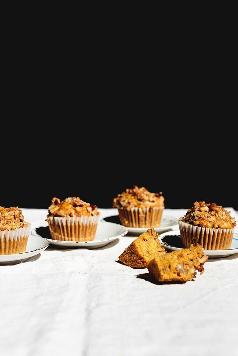Una mesa está cubierta con un mantel blanco.En la mesa hay cuatro Magdalenas de zanahoria y nueces enteras. La quinta está cortada. Se ve muy esponjosa.