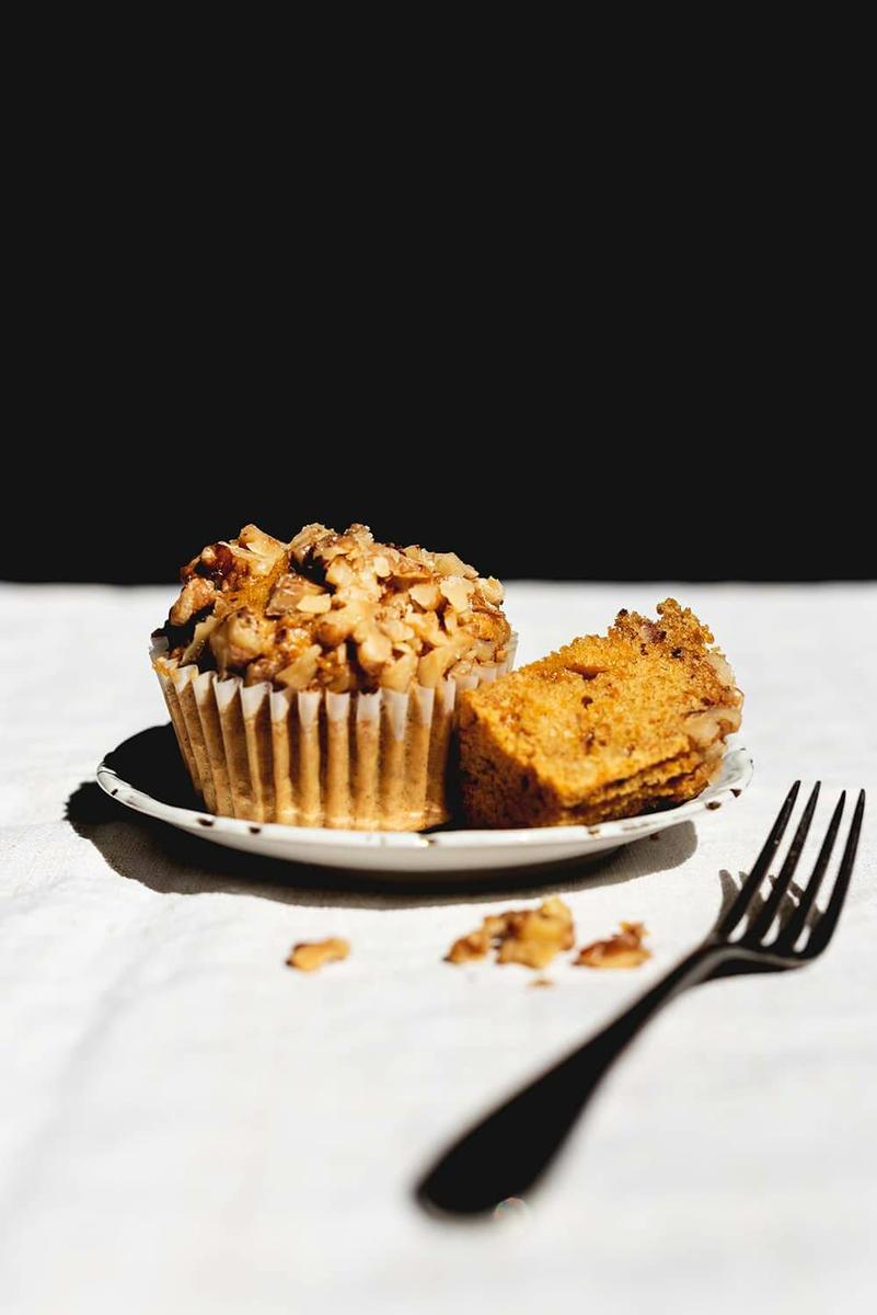 Una mesa está cubierta con un mantel blanco.En la mesa hay un plato . En el plato hay una magdalena de zanahoria y nueces y otro trozo cortado. Al lado del plato hay un tenedor.