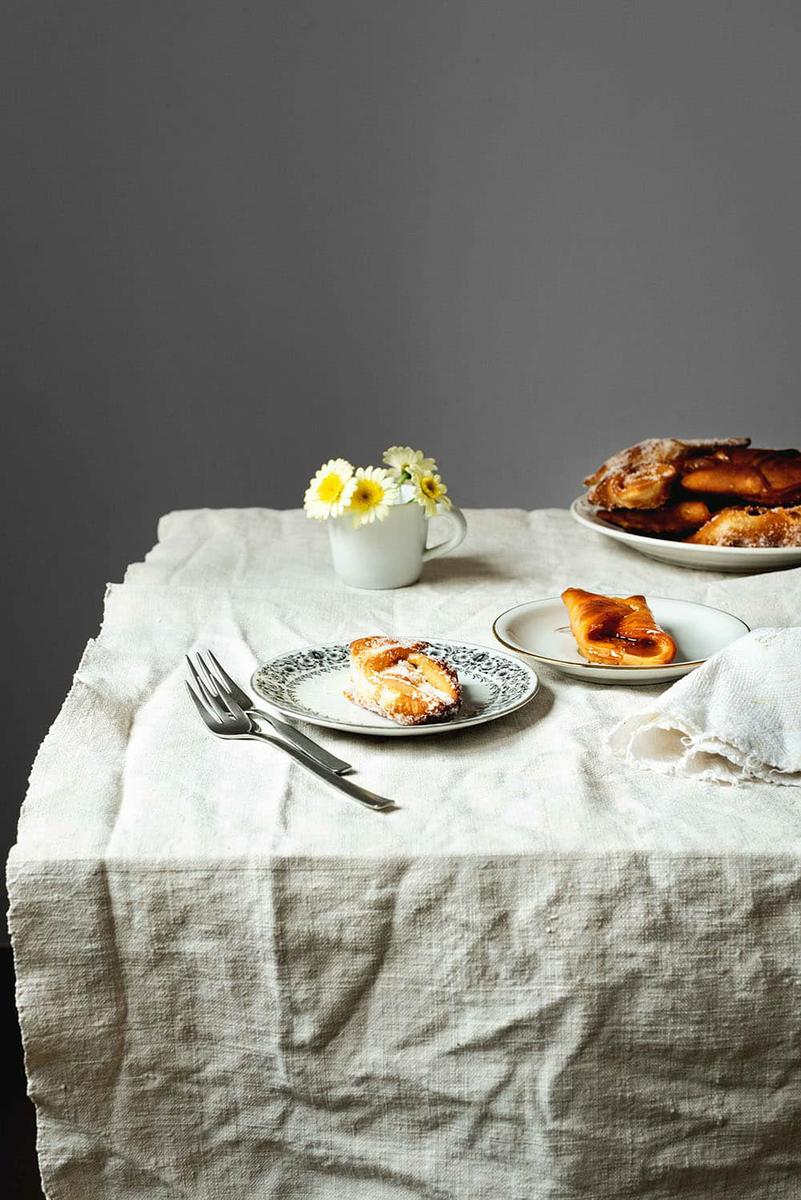 En una mesa hay un mantel blanco. En la mesa hay un plato lleno de pestiños crujientes con miel de la abuela. Al lado de este plato hay flores en un vaso blanco. En el centro de la mesa hay dos platos en cada una hay pestiños crujientes de la abuela.