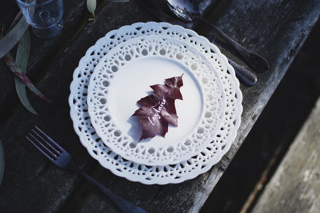 La mesa situada en pleno bosque acoge un banquete en petit comité. La vajilla, las hojas con nombres de árboles escritos a mano y las flores en botellas de cristal aportan delicadeza y calidez a la decoración. Mientras que las hojas secas, los troncos y la mesa de madera natural aportan la esencia propia del otoño. Las frutas de temporada también toman protagonismo en esta celebración tanto en la decoración como en el postre.